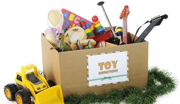 A large corrugated box with a "toy donations" sign filled with assorted toys and surrounded by Christmas garland and a toy bulldozer.© Can Stock Photo / McIninch