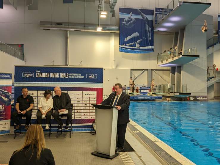 As divers practice in the background, TWEPI CEO Gordon Orr announces the 2024 Canadian Olympic Diving Trials at the Windsor International Aquatic and Training Centre, May 15, 2024. Photo by Mark Brown/WindsorNewsToday.ca.